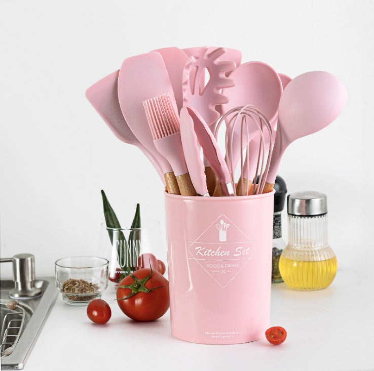 Pink kitchen utensil set in a container on a white surface with tomatoes and a kitchen sink in the background.