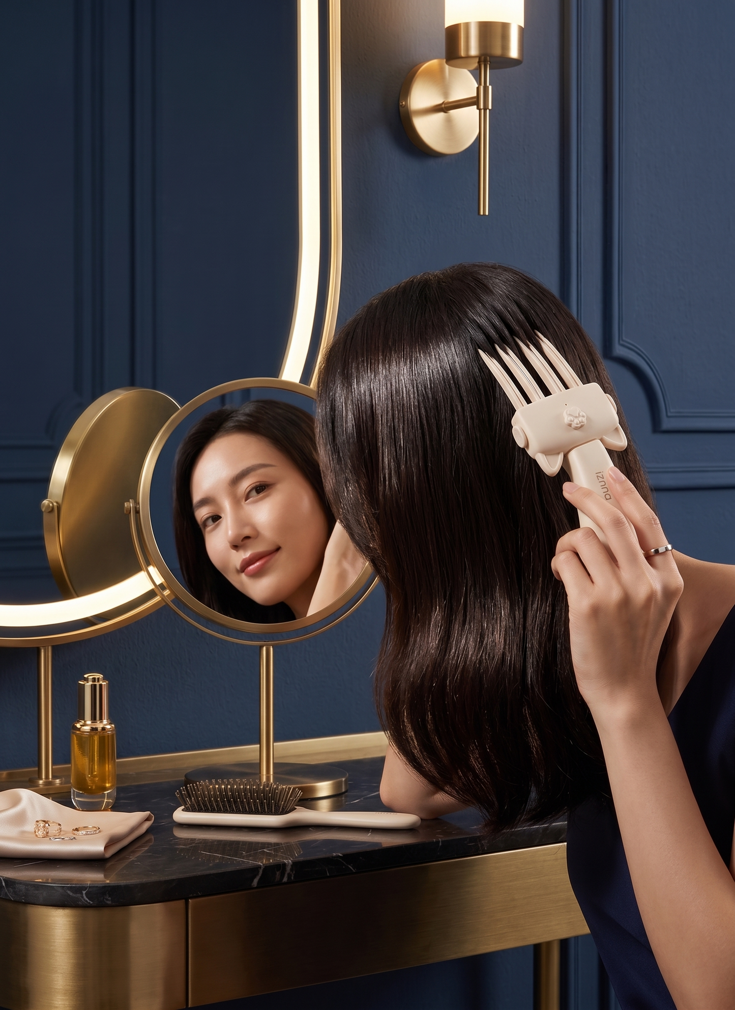 A woman with long dark brown hair is shown from behind in a vanity setting, using a cream-colored hair detangling brush with a decorative elephant head handle. She is looking into a round vanity mirror with a gold stand, set against a dark blue paneled wall with a gold sconce.